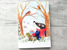 Load image into Gallery viewer, An A6 lined notebook with a drawing of a badger sat with a cup of tea in the countryside in the snow, looking at two robins in the trees
