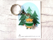 Load image into Gallery viewer, Recycled acrylic brown bear keyring. The bear is wearing a big green jumper which sits just under his nose and he is wearing matching green stripey socks. The keyring is displayed on a small white backing card with a cartoon forest design behind
