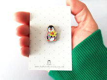 Load image into Gallery viewer, Mini wooden penguin pin badge, on a small white and grey polka dot backing card. The penguin is wearing a pastel rainbow stripe jumper and is holding a daffodil