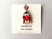 Load image into Gallery viewer, A clear recycled acrylic penguin keyring. This little penguin is holding a bright red heart which has a smiley face. It is displayed on a square white backing card which has the wording sending love and a penguin.