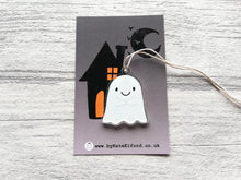 Load image into Gallery viewer, Cute recycled acrylic ghost hanger with a happy face and the hands held together in front. It is displayed on a small backing card that is dark grey with a black house and moon silhouette