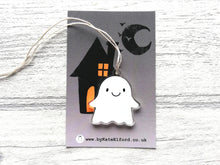 Load image into Gallery viewer, Cute recycled acrylic ghost hanger with a happy face and arms outstretched to the sides. It is displayed on a small backing card that is dark grey with a black house and moon silhouette