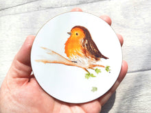 Load image into Gallery viewer, A round coaster, the picture is a watercolour fluffy robin sat on a branch with a few leaves and one leaf falling. The background is plain white. Shown in a hand as a size guide