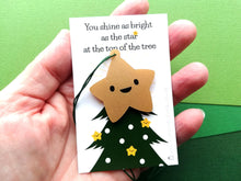 Load image into Gallery viewer, Gold acrylic star decoration with a happy face. The backing card is a picture of the top of a Christmas tree and the wording says you shine as bright as the star at the top of the tree. It is photographed in a hand to show the size