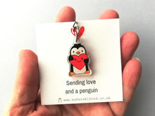 Load image into Gallery viewer, A mini recycled acrylic penguin keyring. The penguin is holding a red heart which has a smiley face. It is displayed on a white backing card with the words sending love and a penguin. It is photographed in my hand to show the size.
