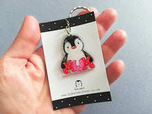 Load image into Gallery viewer, Mum penguin acrylic keyring. The penguin is hugging the word mum in red and pink polka dot letters. Made from recycled acrylic and displayed on a small black and white card. Photo taken in a hand to help show the size 4cm tall