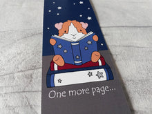 Load image into Gallery viewer, Guinea pig bookmark. A guinea pig sat on a book reading another book. There is a starry sky behind and stars on the book that the guinea pig is reading. The words at the bottom of the bookmark reads one more page