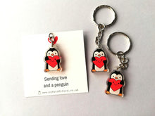 Load image into Gallery viewer, A mini recycled acrylic penguin keyring. The penguin is holding a red heart which has a smiley face. It is displayed on a small square white backing card with the wording sending love and a penguin.
