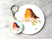 Load image into Gallery viewer, A round glossy coaster and keyring. The image printed on the coaster is a watercolour fluffy robin sat on a branch with a few leaves and one leaf falling. The background is plain white. There is a matching keyring next to the coaster made from recycled acrylic.