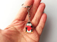 Load image into Gallery viewer, A little penguin keyring in my hand to show the size. Penguin is holding a big red heart which has a happy smiley face.