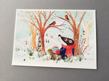 Load image into Gallery viewer, Badger and robins winter countryside print. It measures seven by five inches which is a standard size to make it easy to frame.
The badger is sat under a tree drinking a cup of tea talking to the robins. It is a snowy, hilly scene behind.