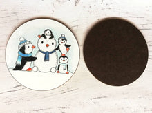 Load image into Gallery viewer, Penguin coaster back and front. Four penguins playing in the snow, building a snow penguin. The penguins are wearing blue hats and scarves
