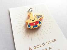 Load image into Gallery viewer, Gold acrylic star, a gold star for a super star stitch marker. The star has a smile and is wearing a blue cape and red pants, holding a flag saying super
