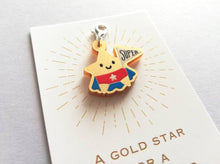 Load image into Gallery viewer, Gold acrylic star, a gold star for a super star stitch marker. The star has a smile and is wearing a blue cape and red pants, holding a flag saying super