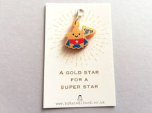 Load image into Gallery viewer, Gold acrylic star, a gold star for a super star stitch marker. The star has a smile and is wearing a blue cape and red pants, holding a flag saying super