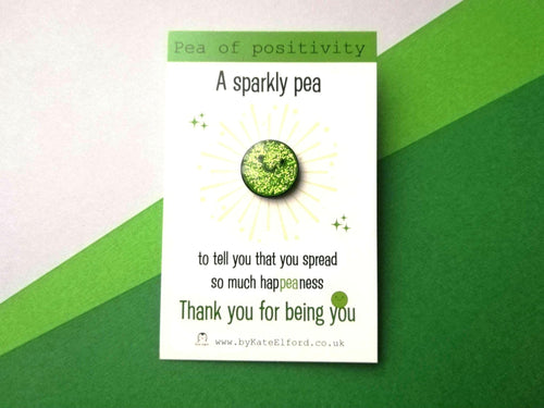 A sparkly green pea enamel pin. It is a round pea with a smiley face. The backing card reads pea of positivity. A, sparkly pea to tell you that you spread so much happiness. Thank you for being you