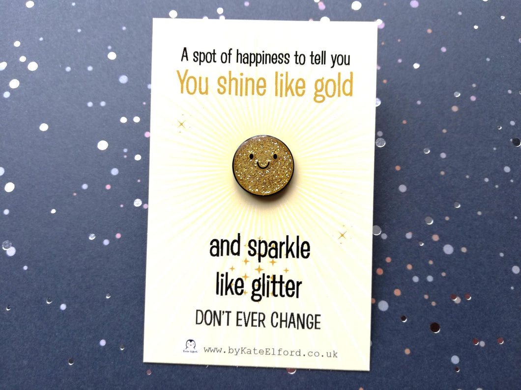 A mini gold glitter round enamel pin with a happysmiling face. It is on a rectangular backing card which has the wording a spot of happiness to tell you you shine like gold and sparkle like glitter. Don't ever change. Designed by Kate Elford