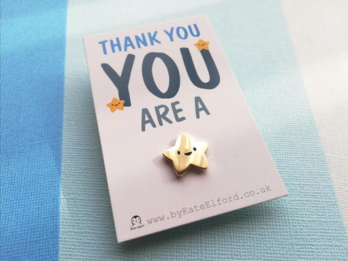 You are a star mini enamel pin. Tiny gold star enamel pin. The star is five pointed and has a happy face. It is on a mini white rectangular backing card with the wording thank you you are a at the top and the star pin is attached at the bottom
