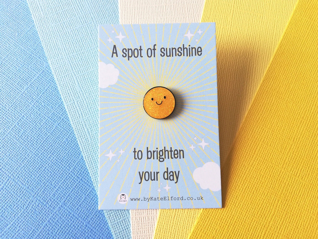 A mini round enamel pin. It is yellow glitter colour with a happy face. The words on the blue backing card read a spot of sunshine to brighten your day