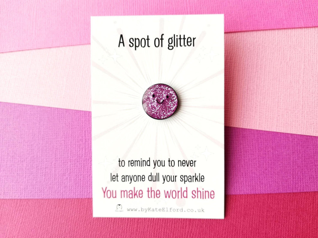 A pink spot of glitter enamel pin with a happy face. The words on the card reads a spot of glitter to remind you to never let anyone dull your sparkle. You make the world shine.