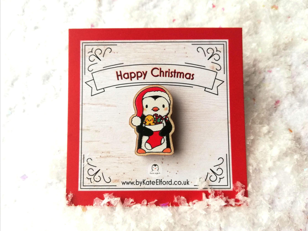 Mini wooden Christmas pin. Penguin wearing a Santa hat and holding a Christmas stocking. The wording on the backing card reads Happy Christmas