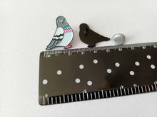 Load image into Gallery viewer, Two pigeon-shaped enamel pins. One pin is a colorful pigeon with grey, blue, pink, and green details. The other is a black silhouette of a pigeon. A ruler shows the pins are approximately 2.5 cm in length.
