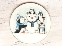 Load image into Gallery viewer, Penguin coaster. Four penguins playing in the snow, building a snow penguin. The penguins are wearing blue hats and scarves

