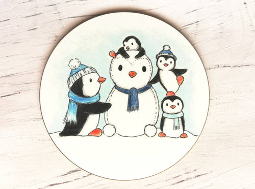 Penguin coaster. Four penguins playing in the snow, building a snow penguin. The penguins are wearing blue hats and scarves