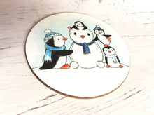 Load image into Gallery viewer, Penguin coaster. Four penguins playing in the snow, building a snow penguin. The penguins are wearing blue hats and scarves
