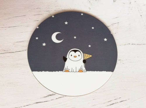 Round coaster. A little penguin dressed as a ghost. There are stars and the moon in the sky behind. Penguin is holding a flag that says Boo