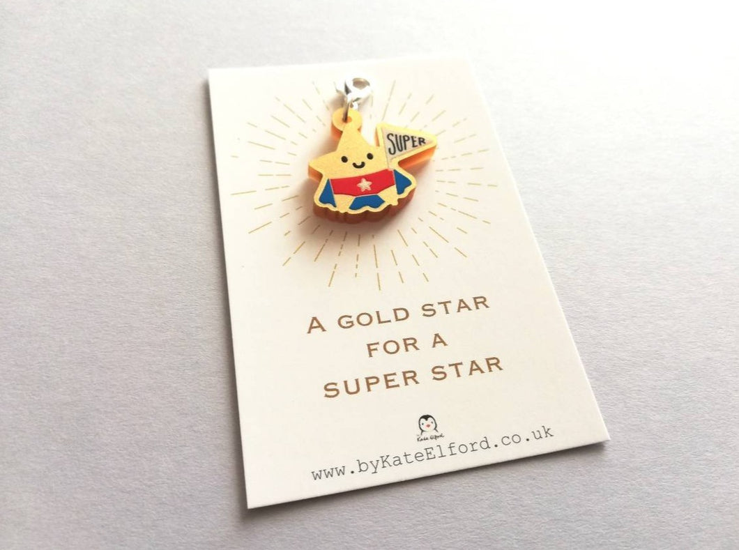 Gold acrylic star, a gold star for a super star stitch marker. The star has a smile and is wearing a blue cape and red pants, holding a flag saying super