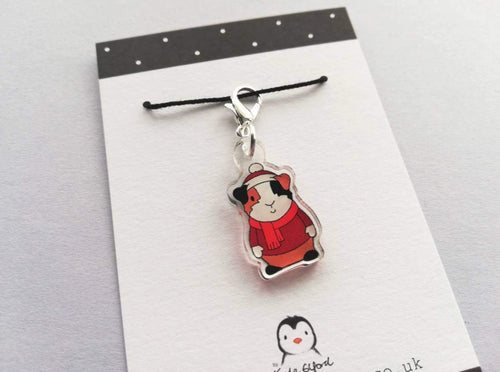 Little clear acrylic guinea pig stitch marker. It is made from recycled acrylic, the guinea pig is wearing a red hat, jumper and scarf